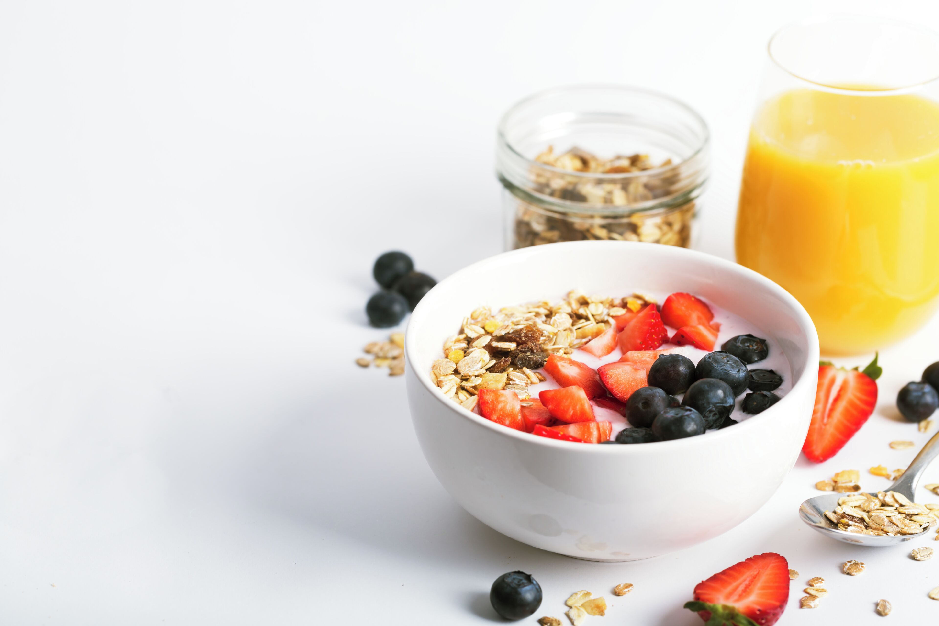 Un tazón de cereales con fresas y arándanos, un vaso de jugo de naranja y un tarro de granola.
