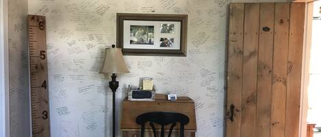 A room with a wall covered in handwritten notes, a framed photo, a desk with a lamp, and a wooden door.