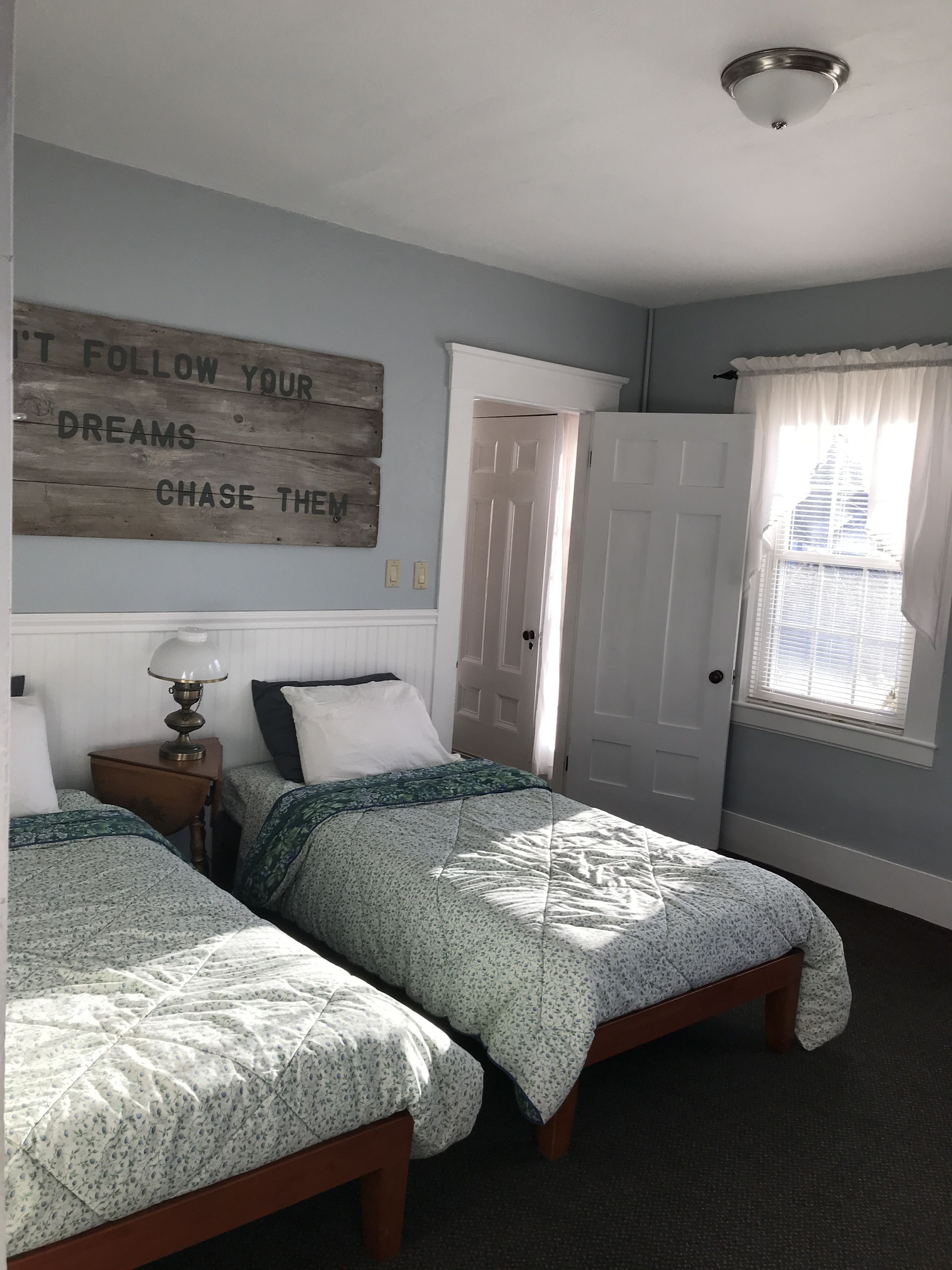 A room with two beds, a motivational quote on the wall, a lamp, and a window with a view of a building.