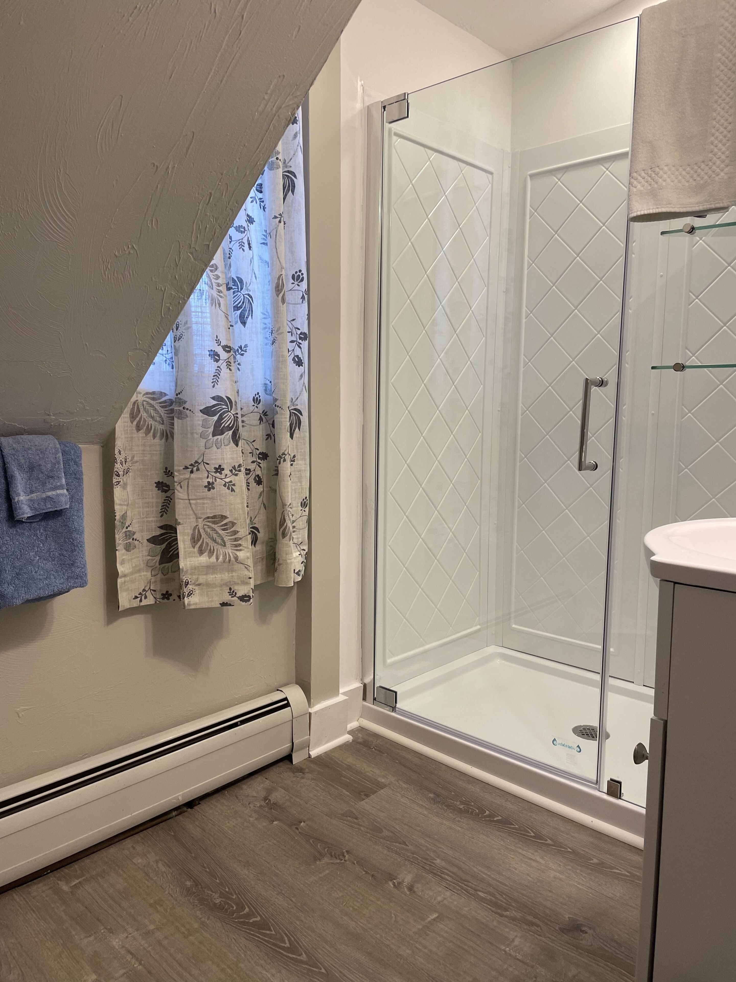 A modern bathroom with a glass shower enclosure, a white tiled shower area, a wooden floor, and a towel hanging on the wall.