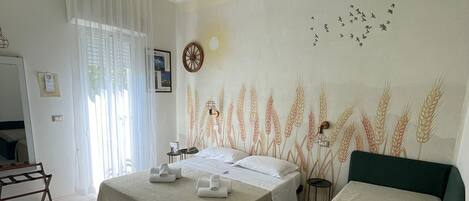 A hotel room with two beds, a large window with sheer curtains, a wall mural of wheat, and a ceiling light fixture.