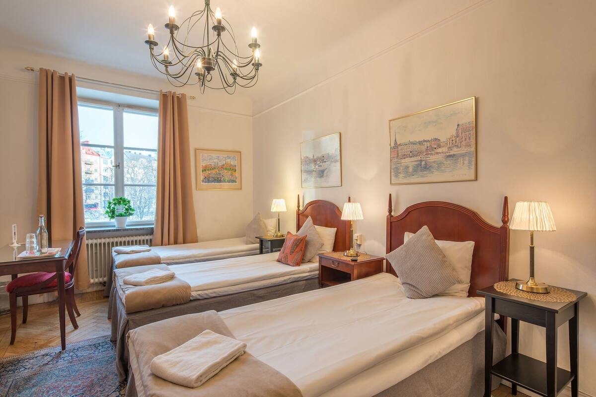 Una habitación de hotel con tres camas, una araña, un escritorio con silla y una ventana con cortinas.