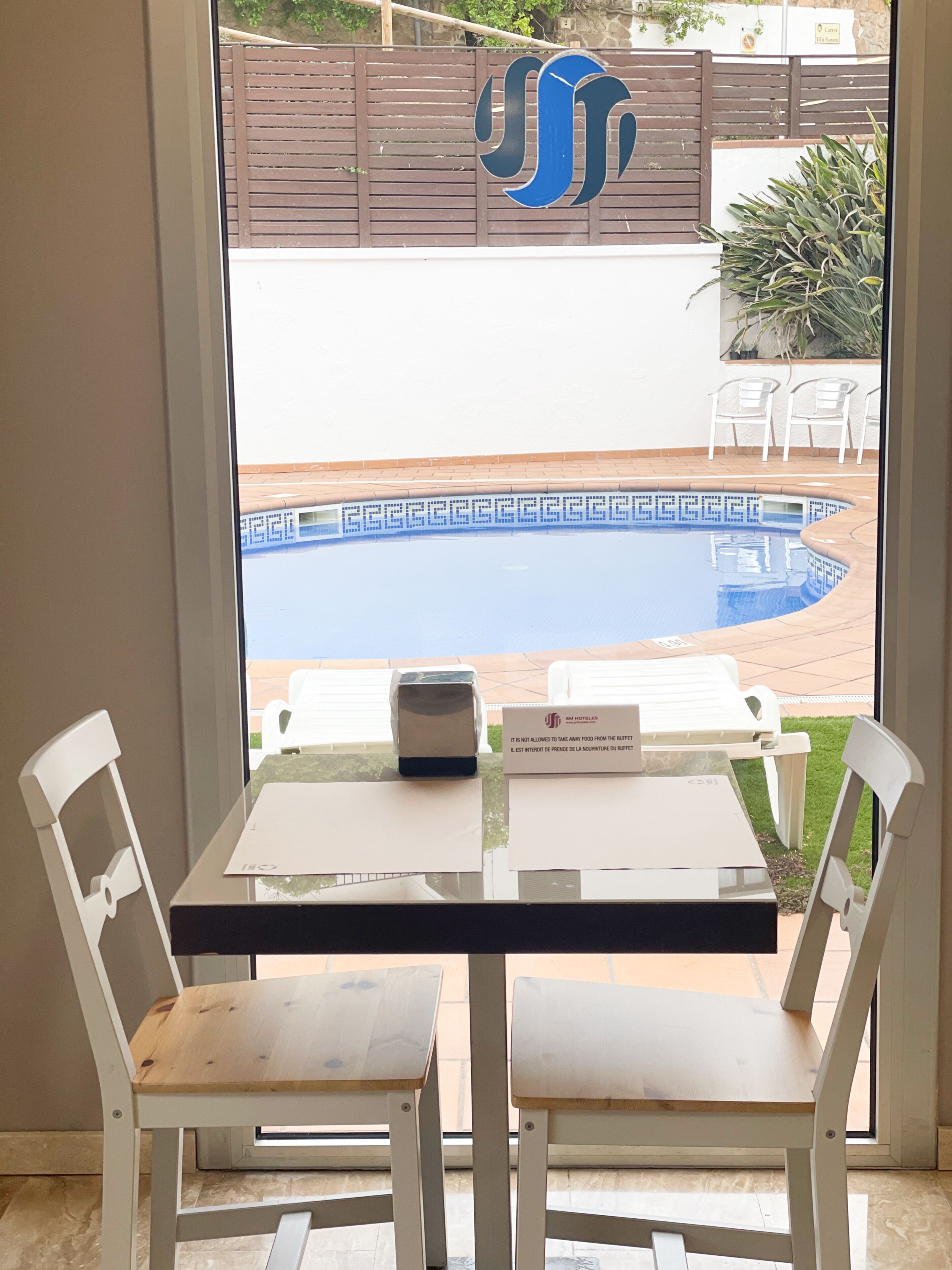 A view of a poolside area with a table and chairs, a sign with a logo, and a fence in the background.