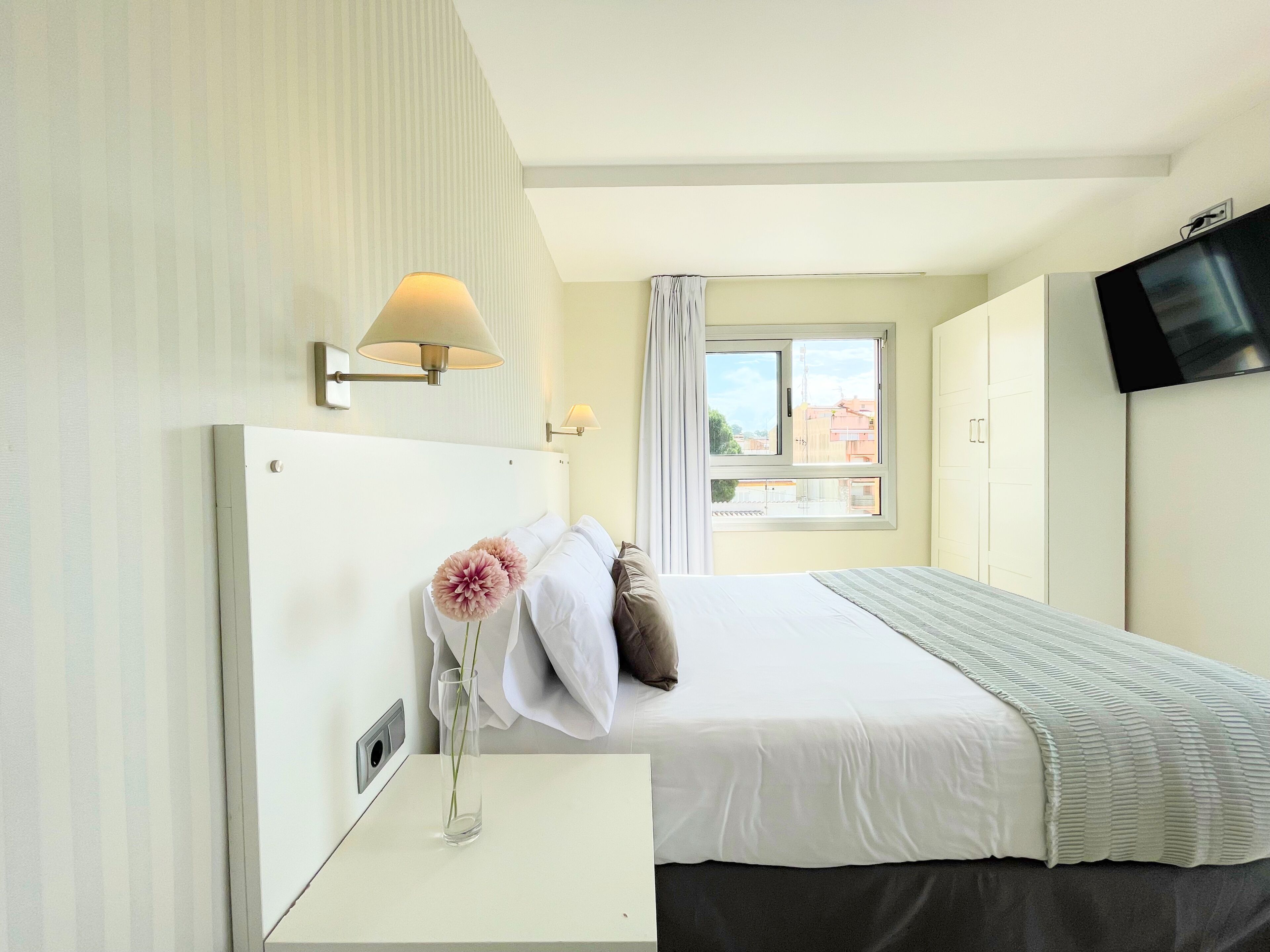 A hotel room with a bed, a bedside table, a lamp, a window with curtains, and a television mounted on the wall.