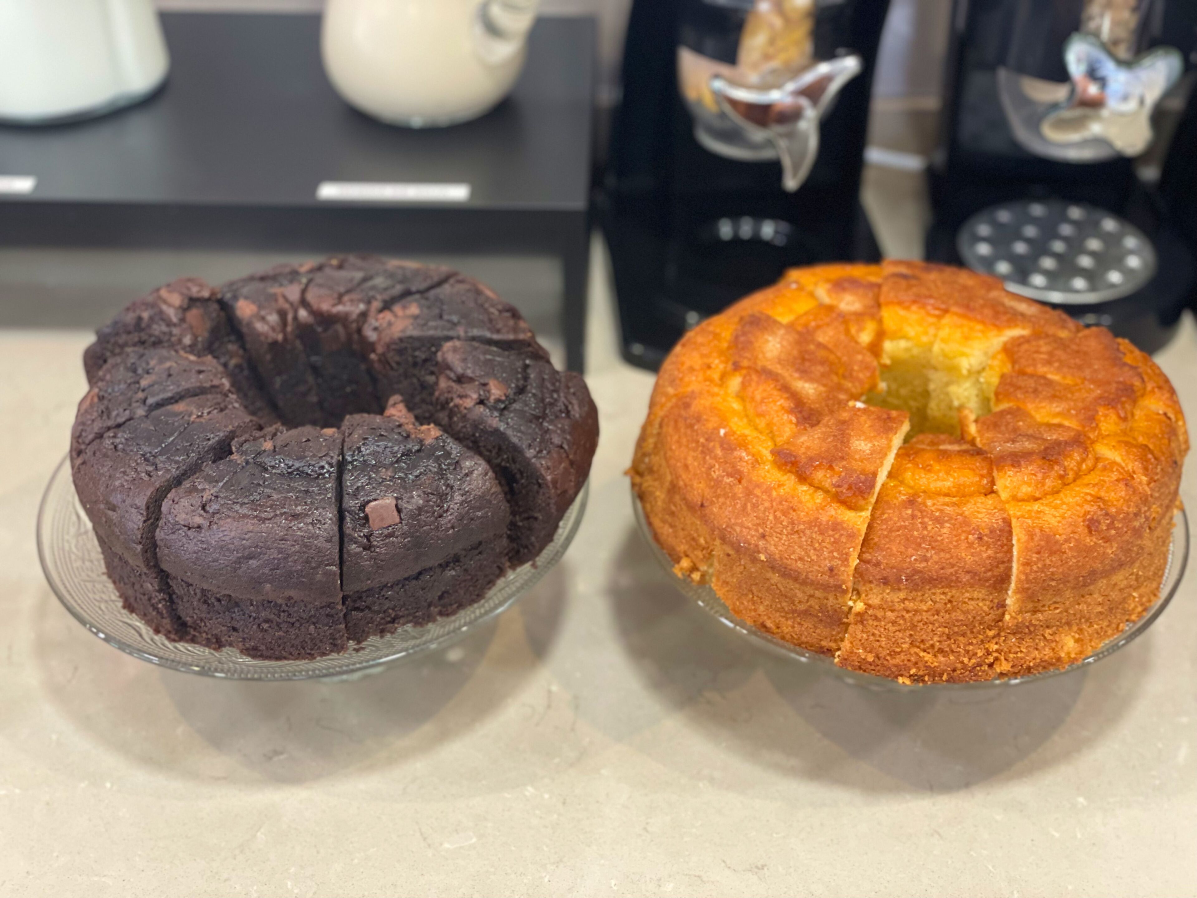Two cakes on glass plates, one chocolate and one yellow, with a coffee machine and a kettle in the background.
