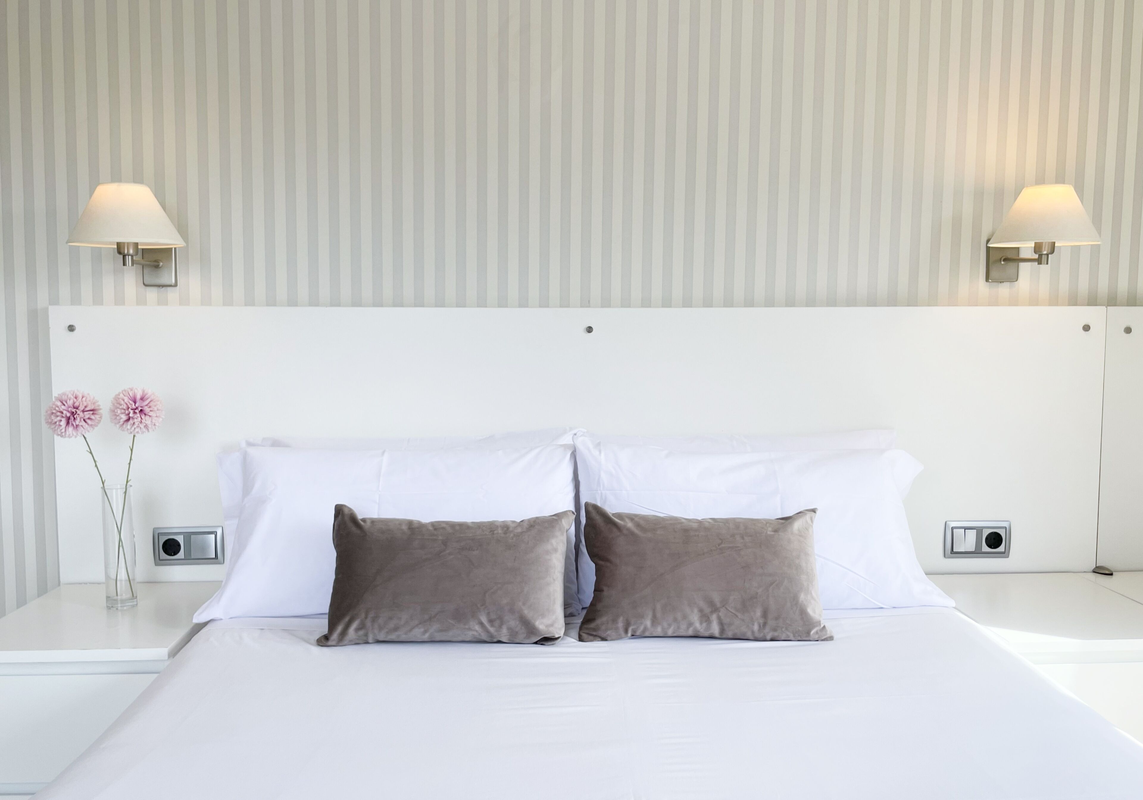 A hotel room with a large bed, two pillows, a nightstand with a lamp, and a vase with flowers.