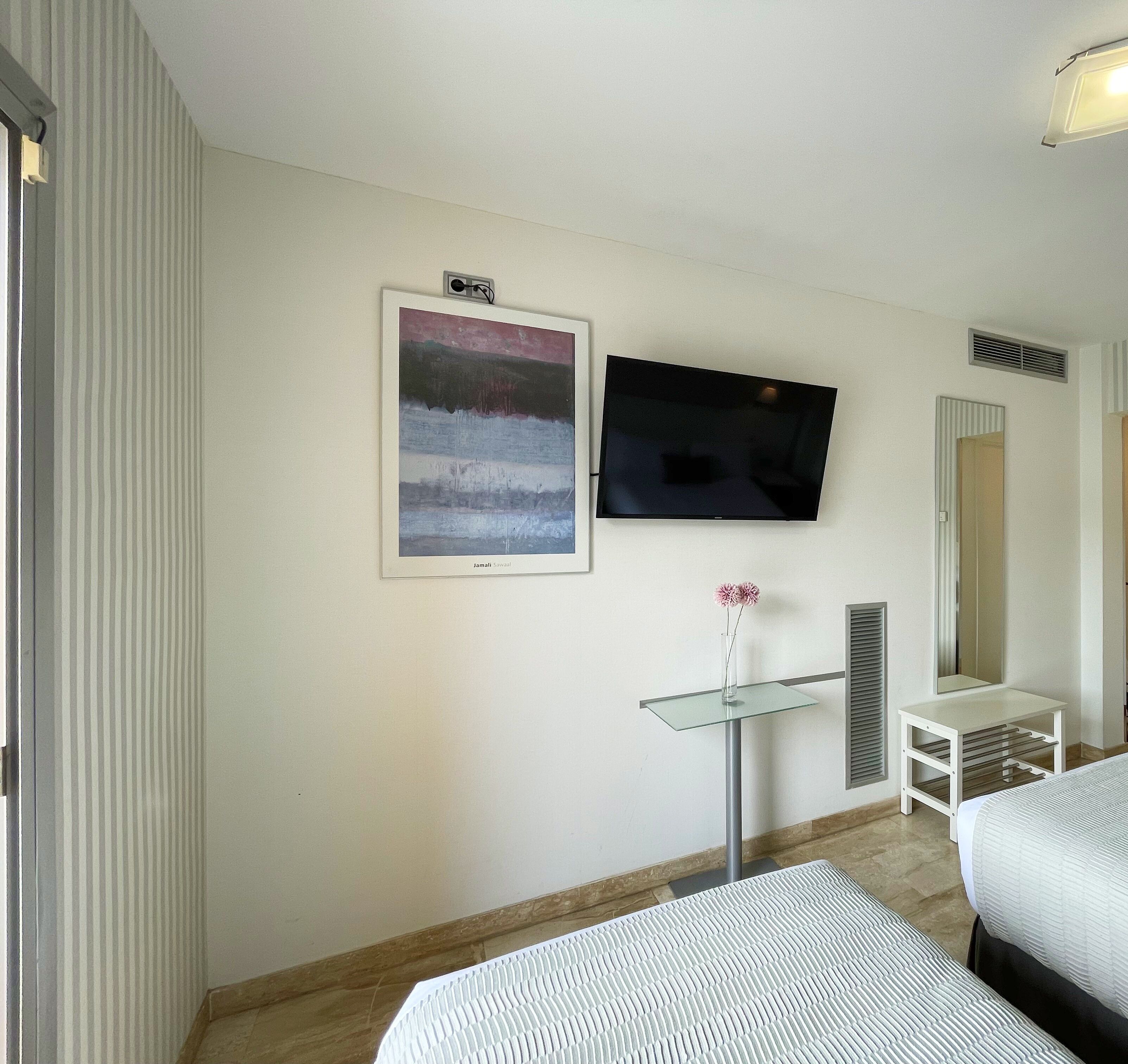 A hotel room with a bed, a TV mounted on the wall, a framed picture, and a small table with a vase.