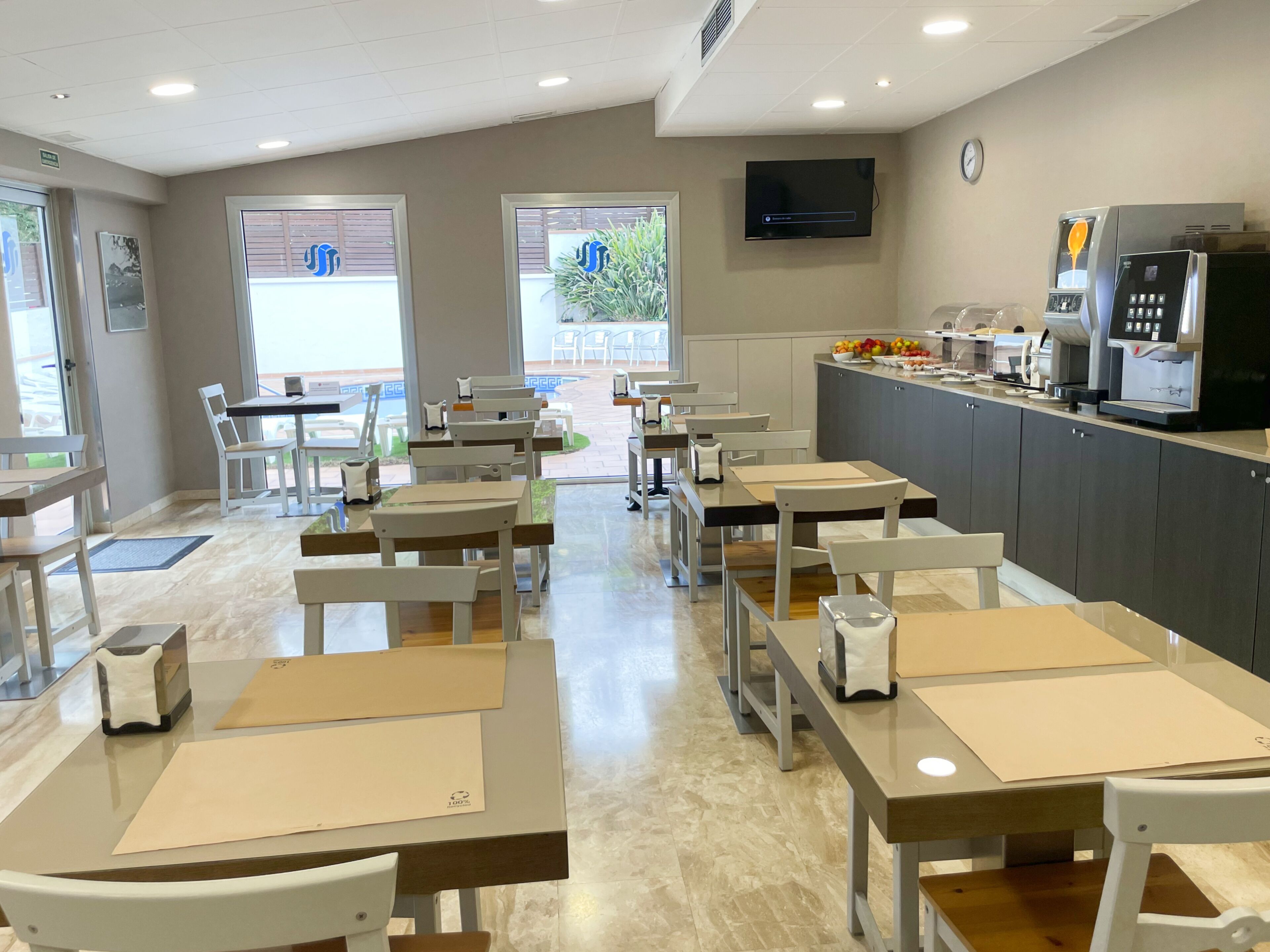 Interior de un restaurante con mesas, sillas, zona de cocina y un televisor en la pared.