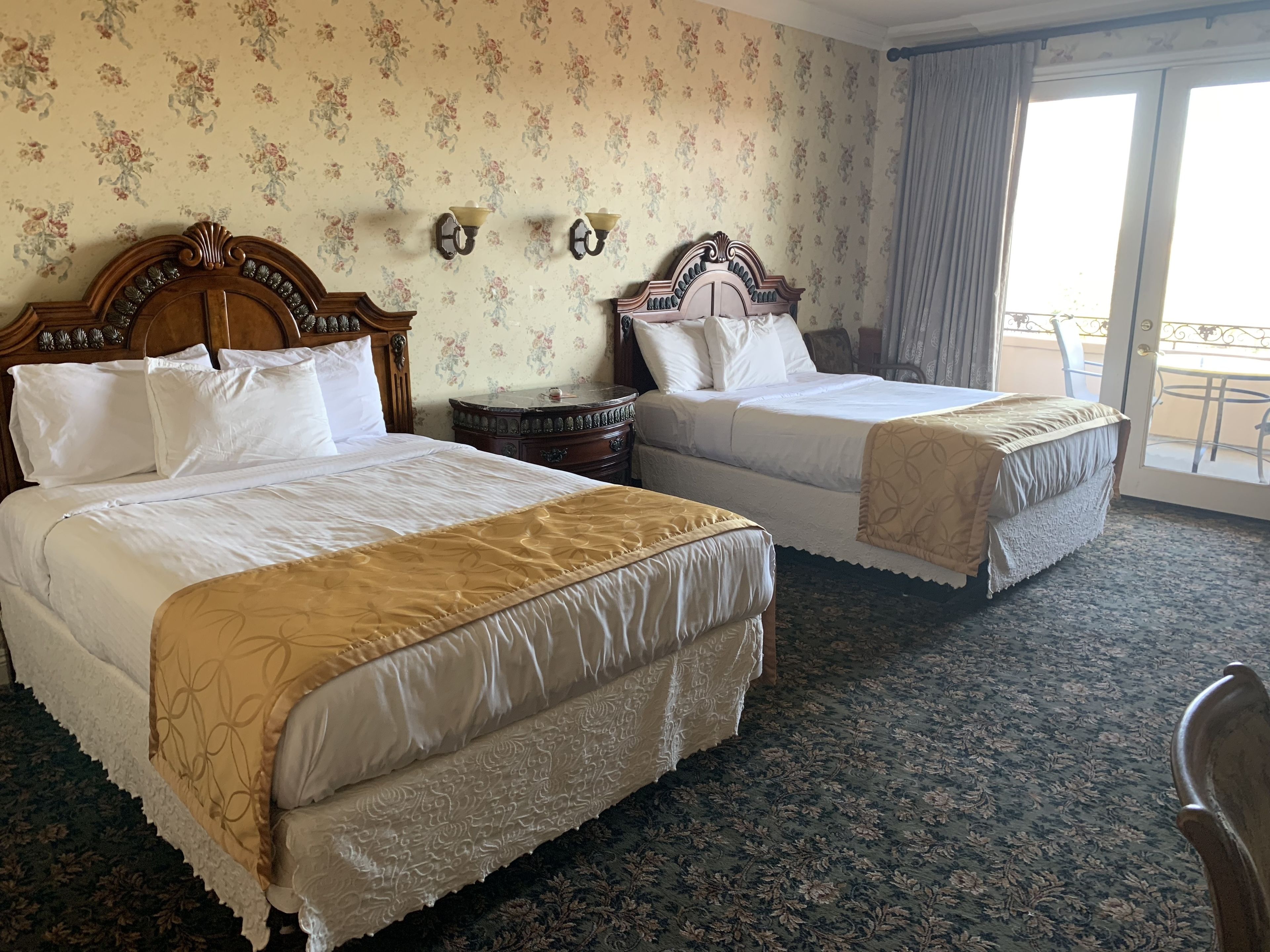 A hotel room with two beds, floral wallpaper, a wooden headboard, a small round table, and a balcony with a railing.