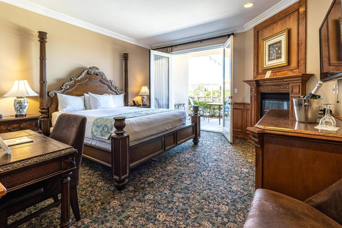 Habitación de hotel con una cama grande, un escritorio con lámpara, una silla, chimenea y un balcón con vistas.