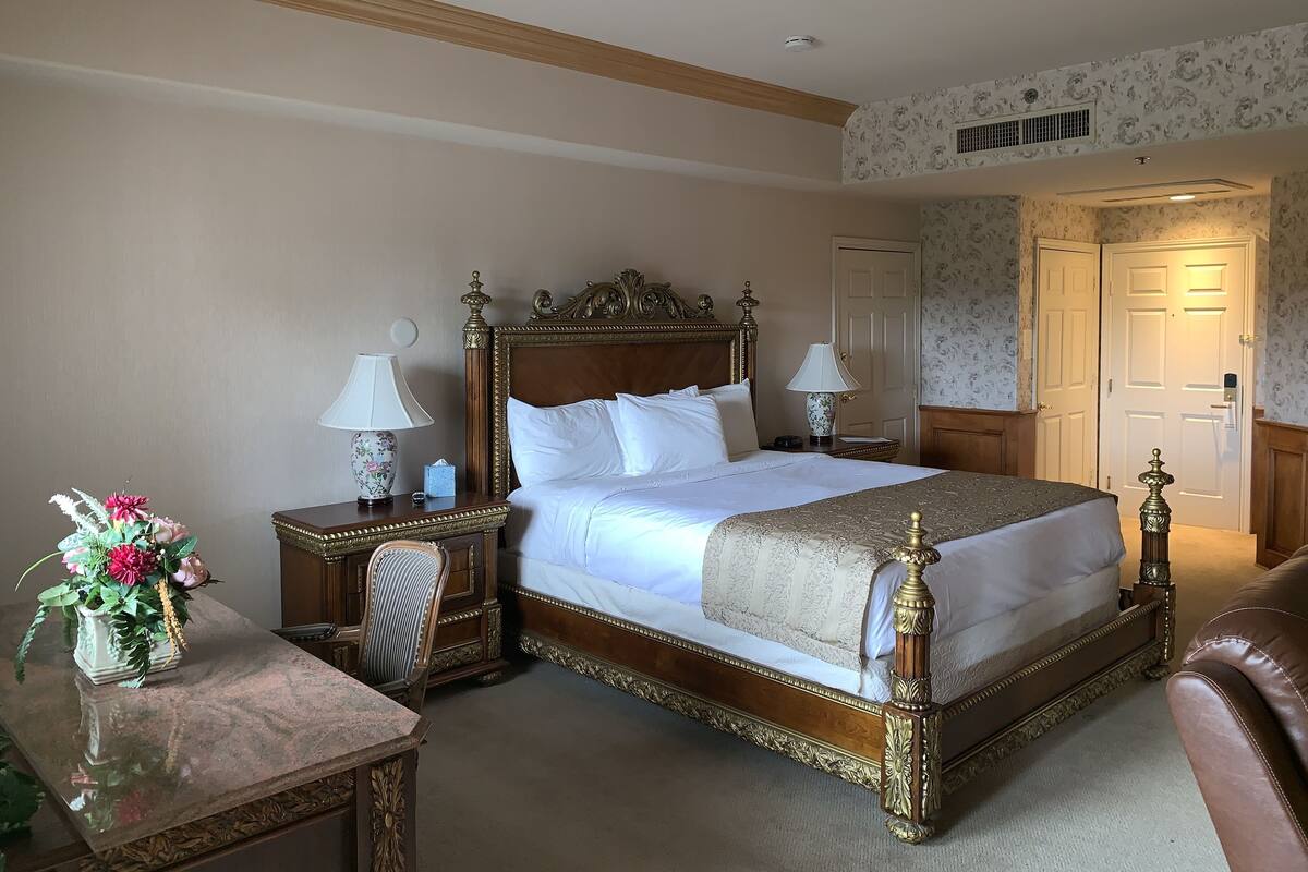 Una cama grande y elaborada con cabecera de madera, dos mesitas de noche con lámparas, una silla y una pequeña mesa con un jarrón de flores.