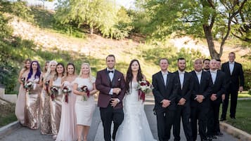 Eine Gruppe von Personen in festlicher Kleidung steht auf einem Weg, im Hintergrund Bäume und Grünpflanzen.