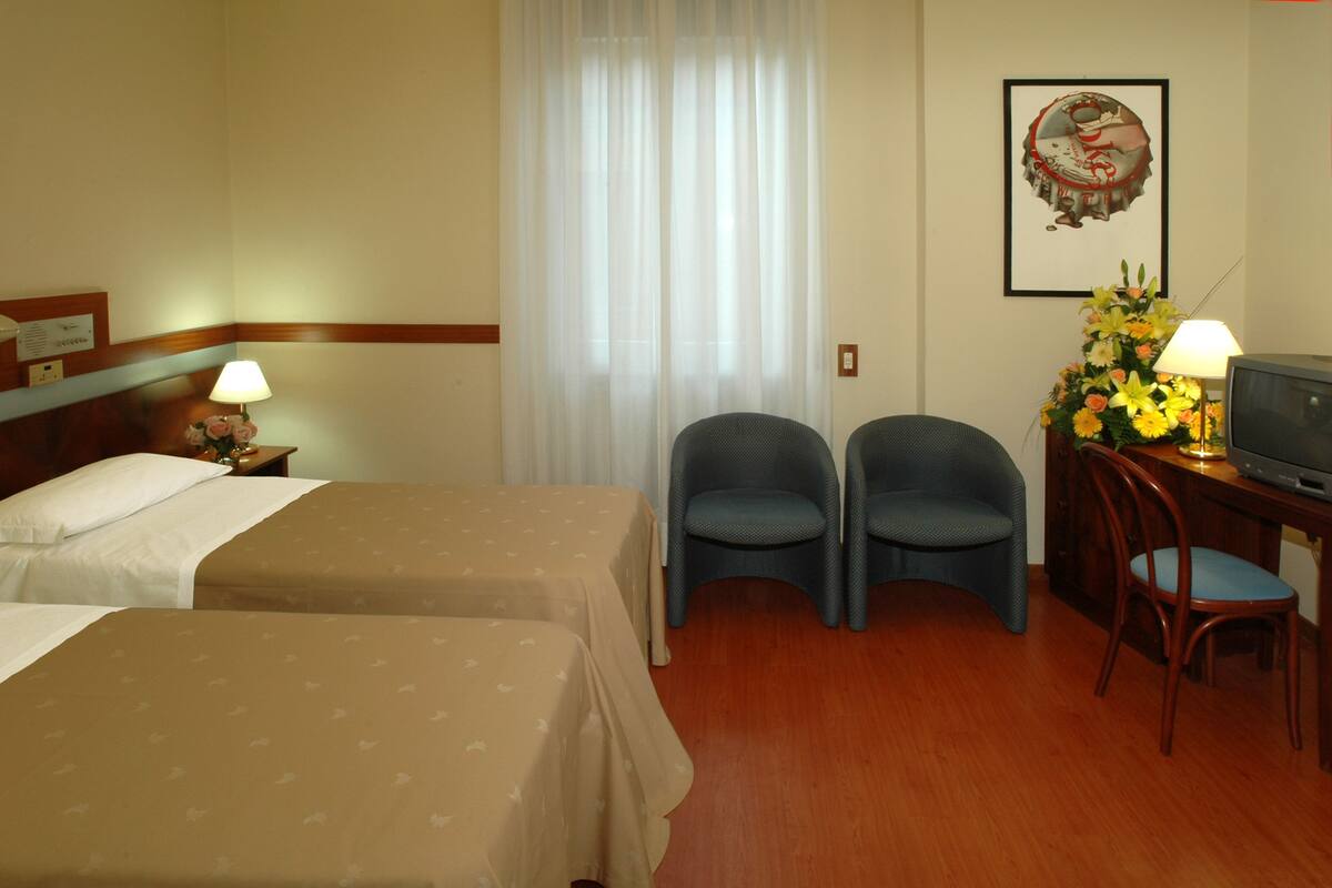 Ein Hotelzimmer mit zwei Betten, einem Schreibtisch mit Fernseher, zwei Sesseln, einer Schreibtischlampe und einer Blumenkomposition.
