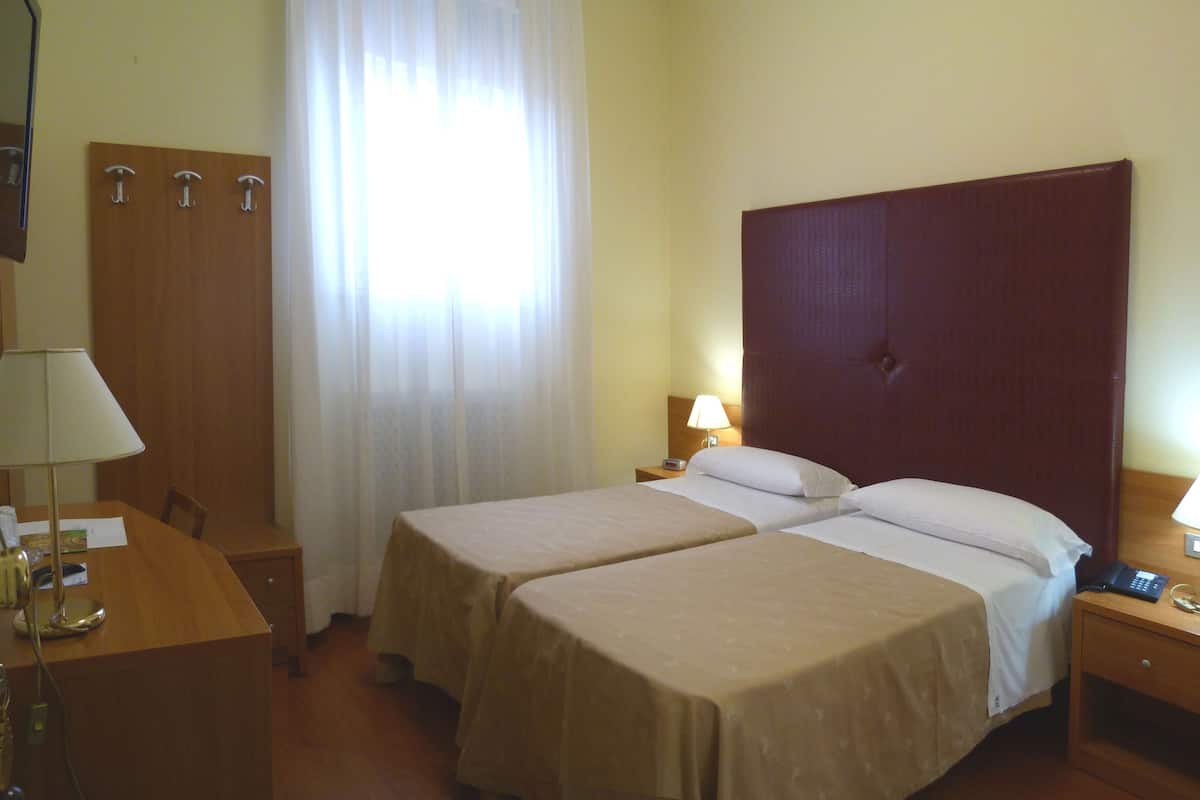 Ein Hotelzimmer mit zwei Betten, einem Schreibtisch, einem Stuhl, einem Spiegel und einem Fenster mit Vorhängen.
