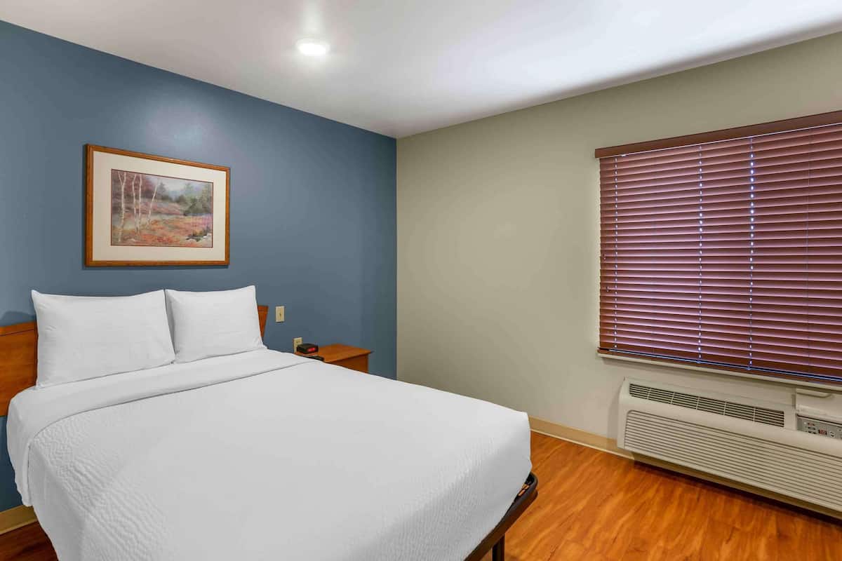 Una cama bien tendida con sábanas blancas, un cuadro en la pared, una mesita de noche con una lámpara y una ventana con persianas.