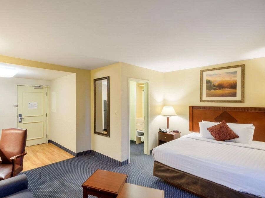 A hotel room with a bed, a chair, a small table, a nightstand with a lamp, and a painting on the wall.