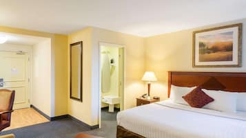 A hotel room with a bed, a chair, a desk, a nightstand, a lamp, a painting, and a bathroom with a shower and toilet.