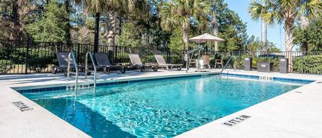 A rectangular swimming pool with a clear blue water surface, surrounded by a white tiled deck. There are lounge chairs and a covered seating area with a table and chairs.