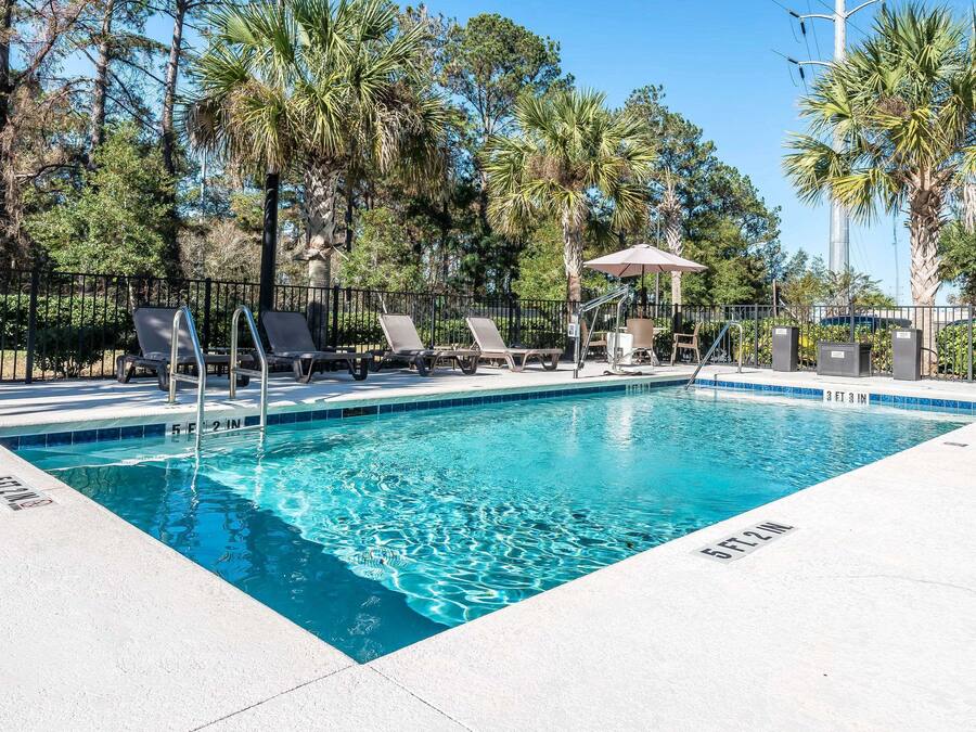 A rectangular swimming pool with a clear blue water surface, surrounded by a white tiled deck. There are lounge chairs and a covered seating area with a table and chairs.