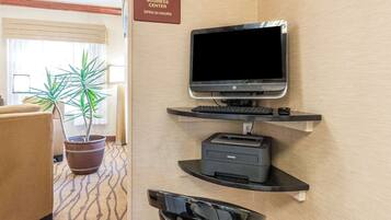 Un espace de réception d’hôtel avec un écran d’ordinateur, un clavier et une imprimante placés sur une étagère fixée au mur. On y trouve également une plante en pot et un panneau indiquant que le centre d’affaires est ouvert 24 heures sur 24.