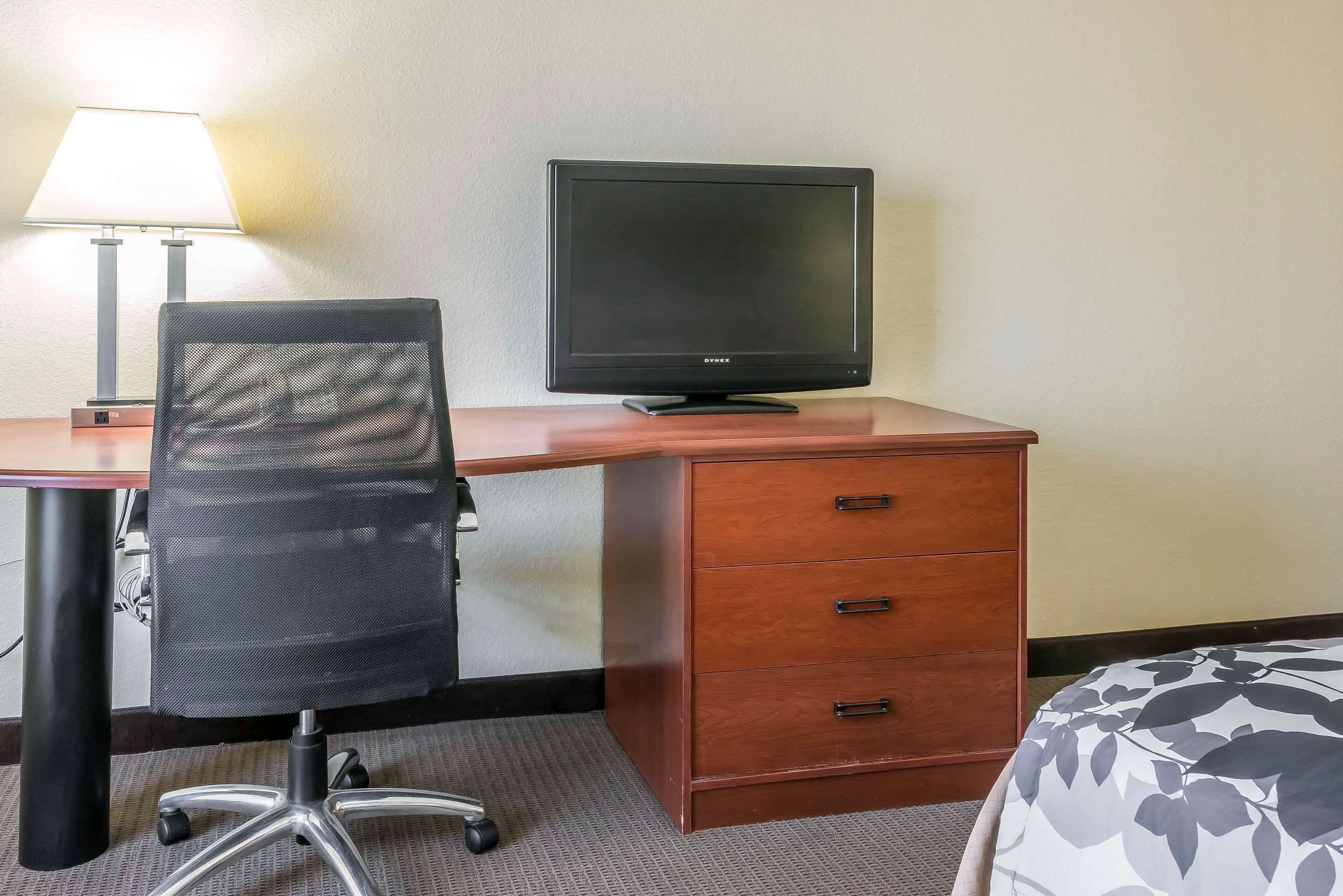 Ein Hotelzimmer mit einem Holztisch, einem schwarzen Bürostuhl, einem Flachbildfernseher und einem Bett mit einem gemusterten Überwurf.
