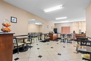 Un comedor de hotel con varias mesas y sillas, una zona de cocina y una cesta de frutas sobre una encimera.