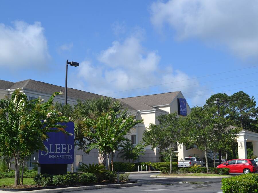 Un bâtiment de la chaîne Sleep Inn & Suites, avec une enseigne, entouré de palmiers et de voitures garées.