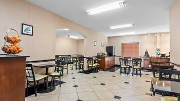 A hotel dining area with multiple tables and chairs, a fruit basket, and a kitchen area in the background.
