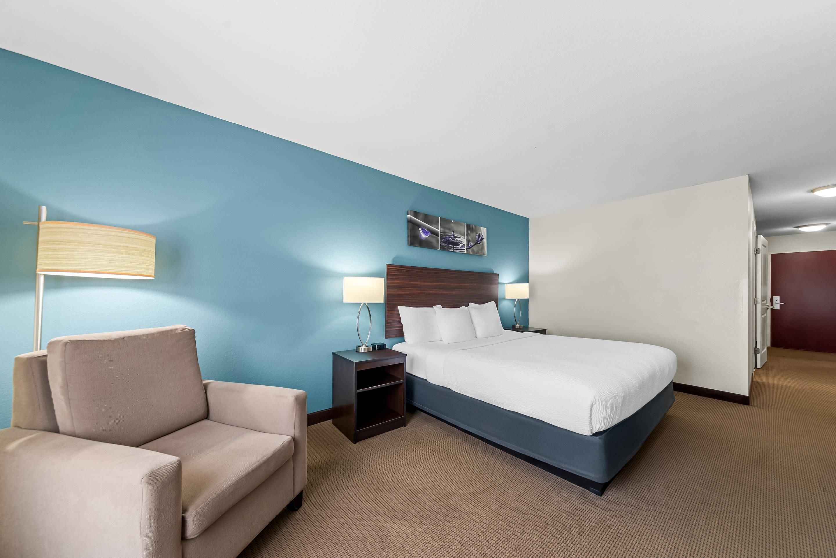 A hotel room with a bed, a chair, a nightstand, a lamp, and a wall art piece.