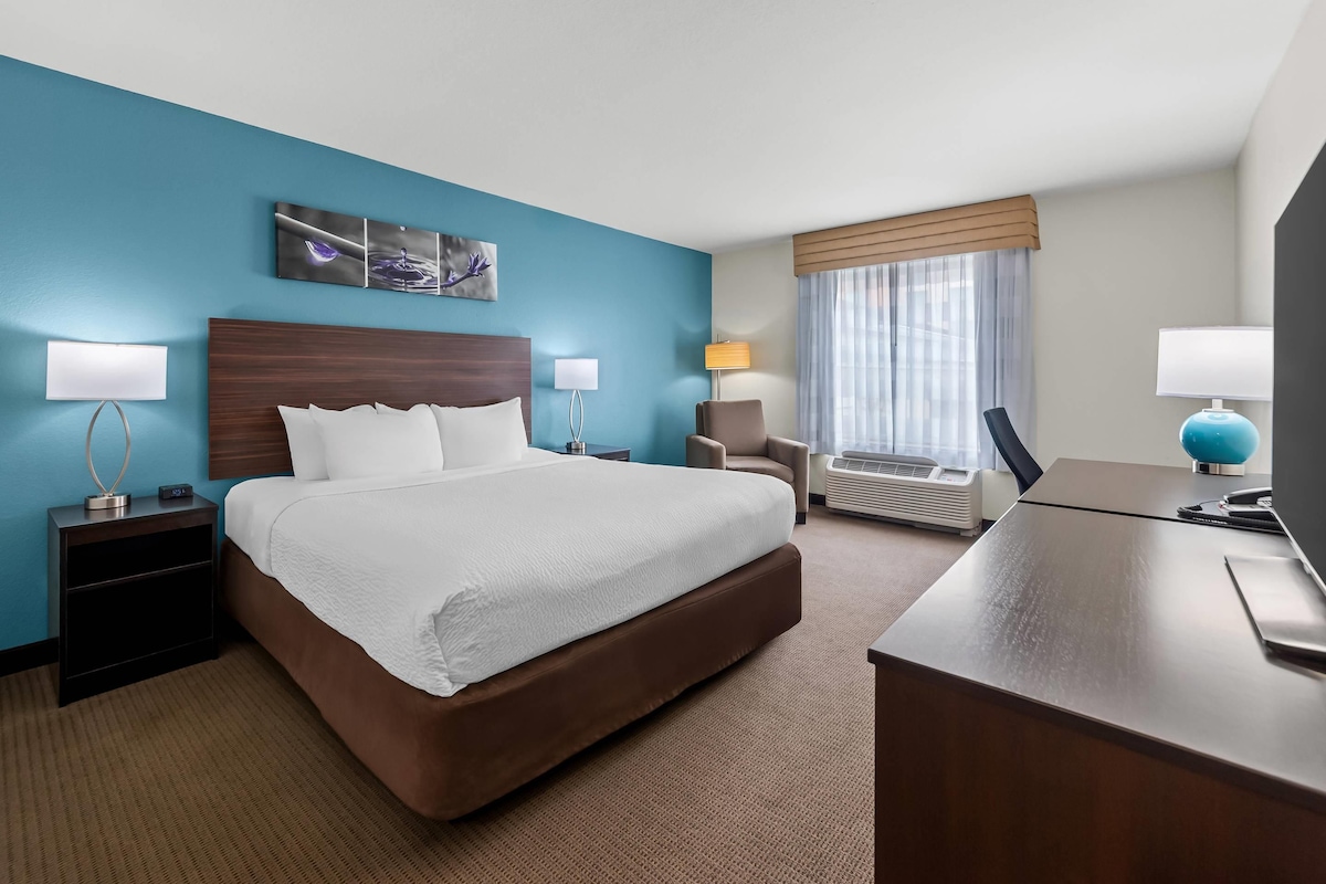 Habitación de hotel con una cama grande, un escritorio, una silla, una mesita de noche, una lámpara y una ventana con cortinas.