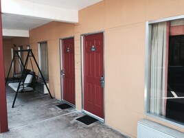Un pasillo con dos puertas rojas, cada una con un número. Hay un sillón mecedor y un escalón que lleva a la puerta.