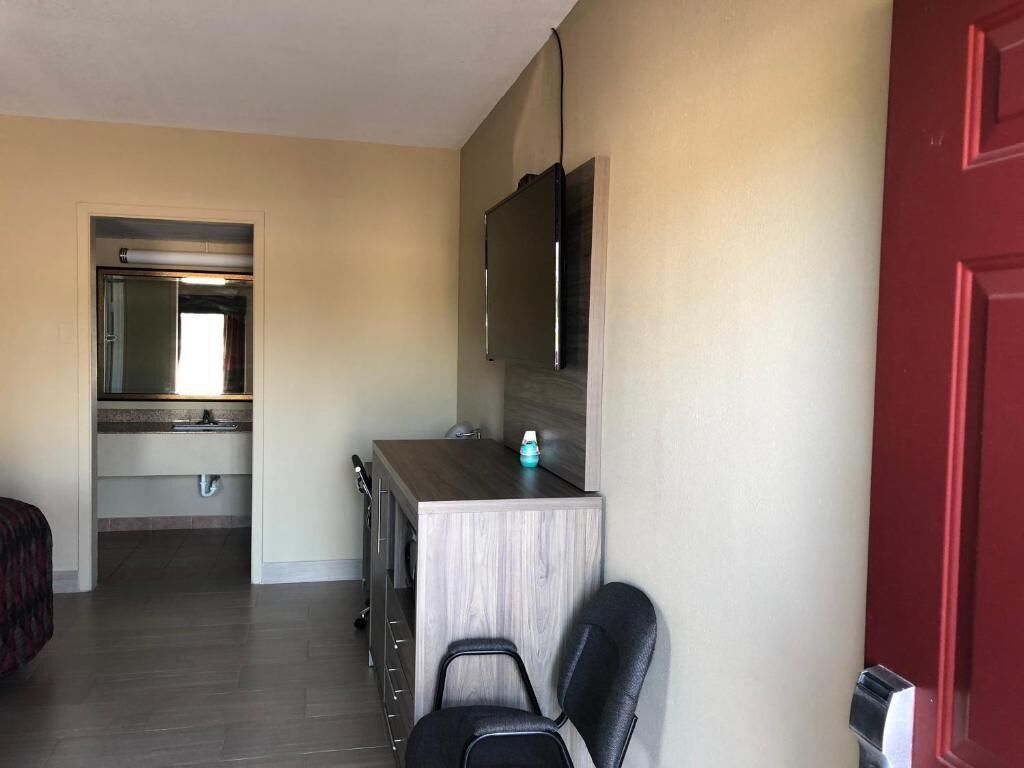 A hotel room with a kitchenette, a bed, a desk with a chair, a TV mounted on the wall, and a red door.