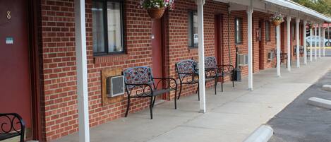 A row of red brick motel rooms with white pillars, outdoor seating, and hanging flower baskets.