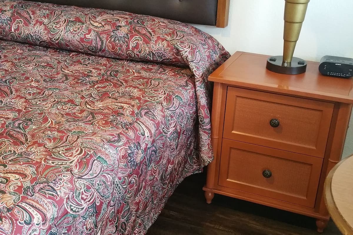 A bedroom with a tufted headboard, a patterned bedspread, a wooden nightstand with drawers, and a bedside lamp.