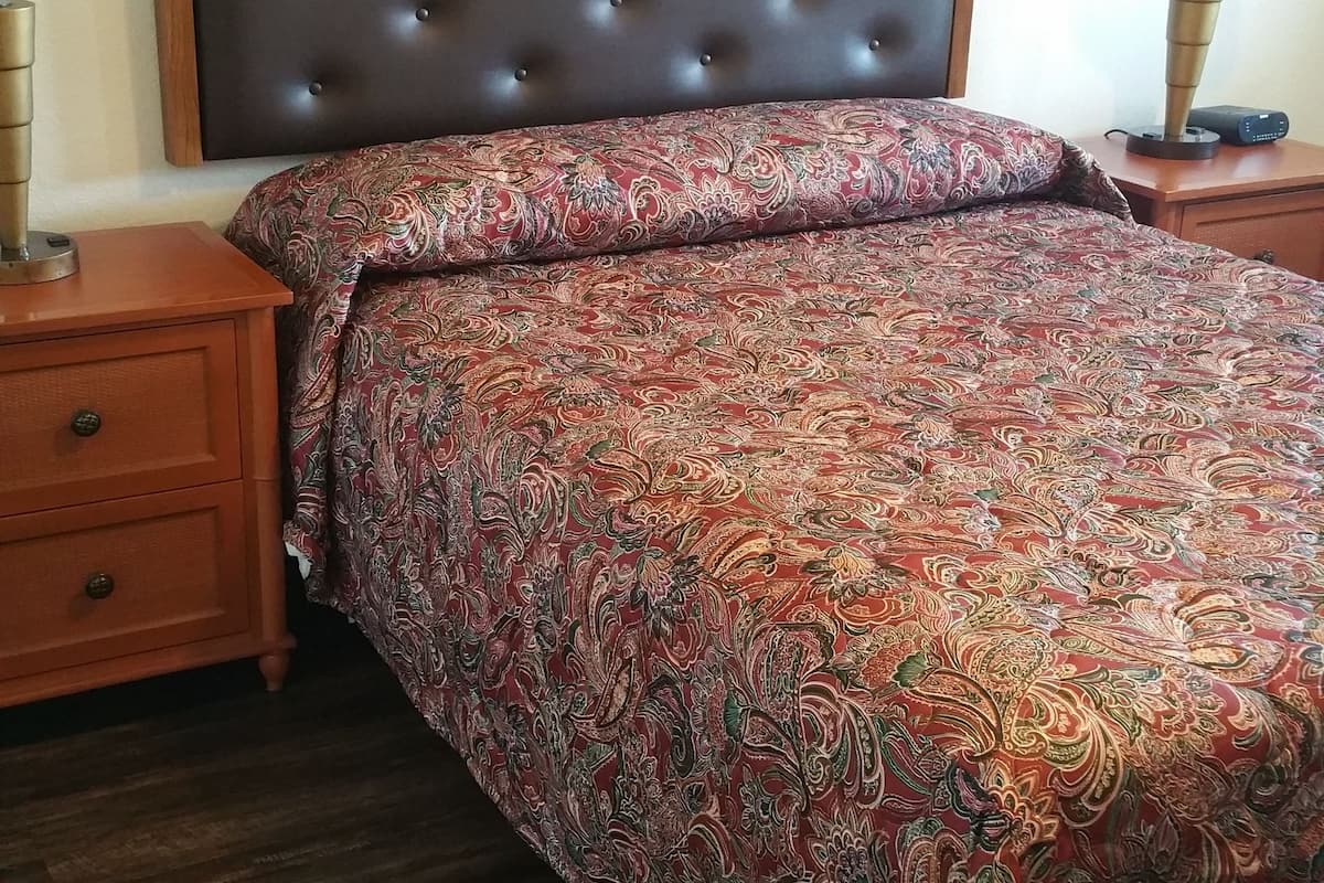 A bedroom with a wooden headboard, a patterned bedspread, a nightstand with a lamp, and a bedside table with a lamp.