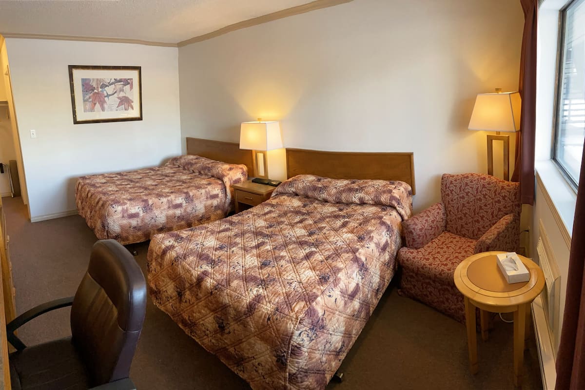 A hotel room with two beds, a chair, a small table, and a lamp.