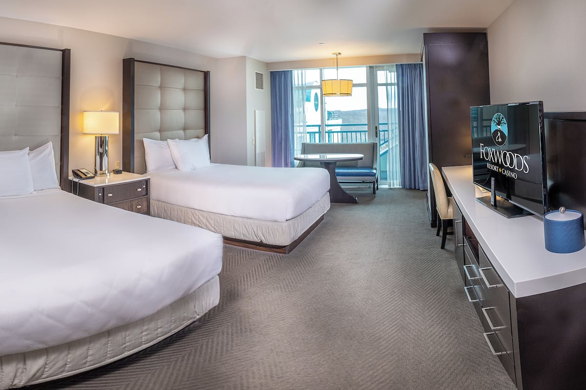 A hotel room with two beds, a desk with a television, and a balcony with a view.