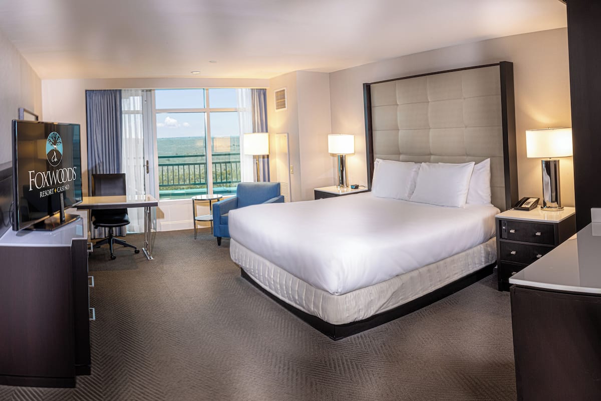 A hotel room with a large bed, a desk, a chair, a TV, and a balcony with a view.