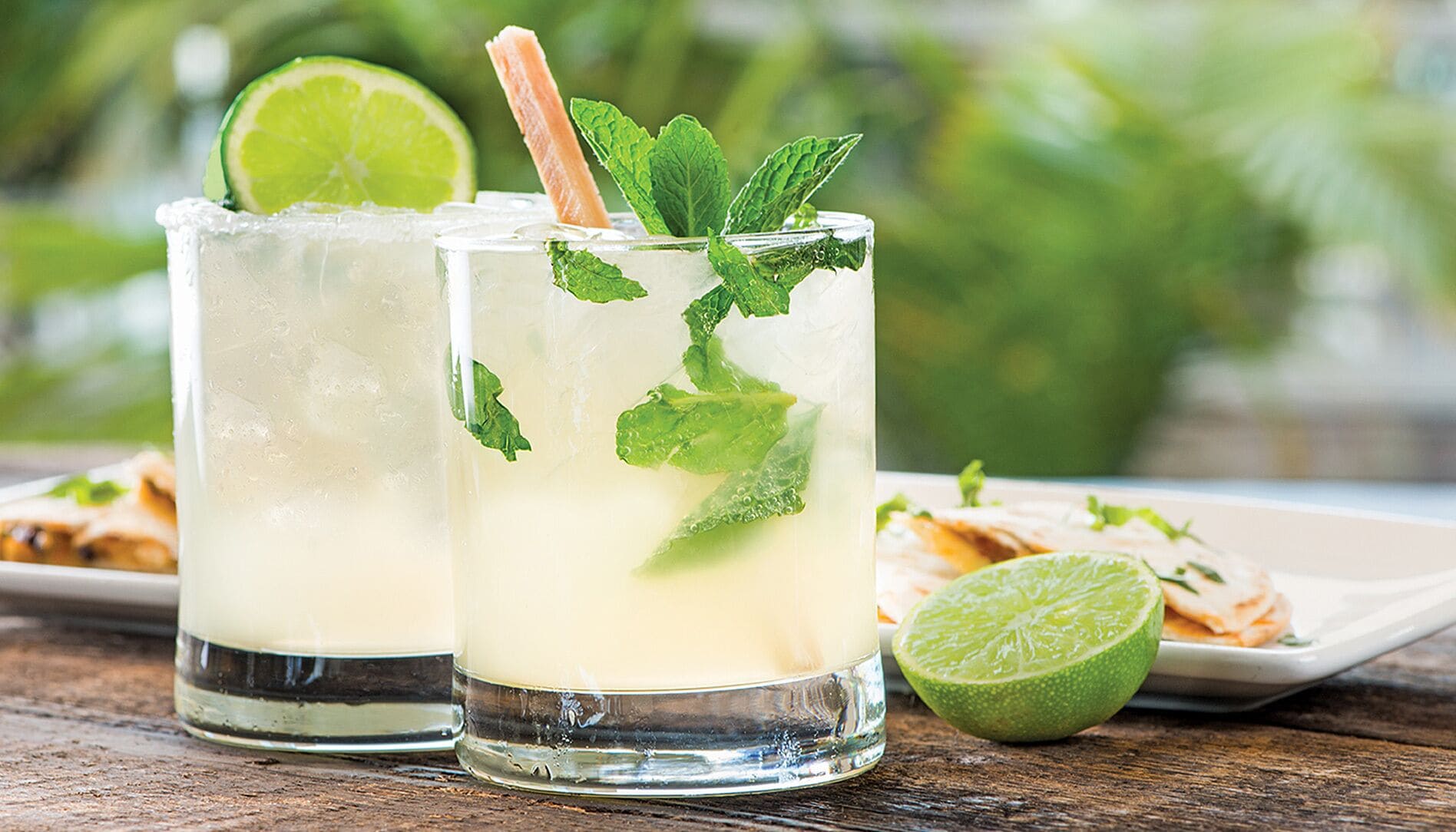 Two glasses of a light-colored beverage with mint leaves and a slice of lime, served on a wooden surface with a plate of food and a lime in the background.