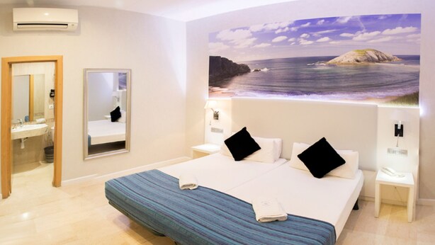 A hotel room with a large bed, a bathroom with a mirror, and a painting of a beach scene on the wall. - Standbyme Ramblas (Barcelona)