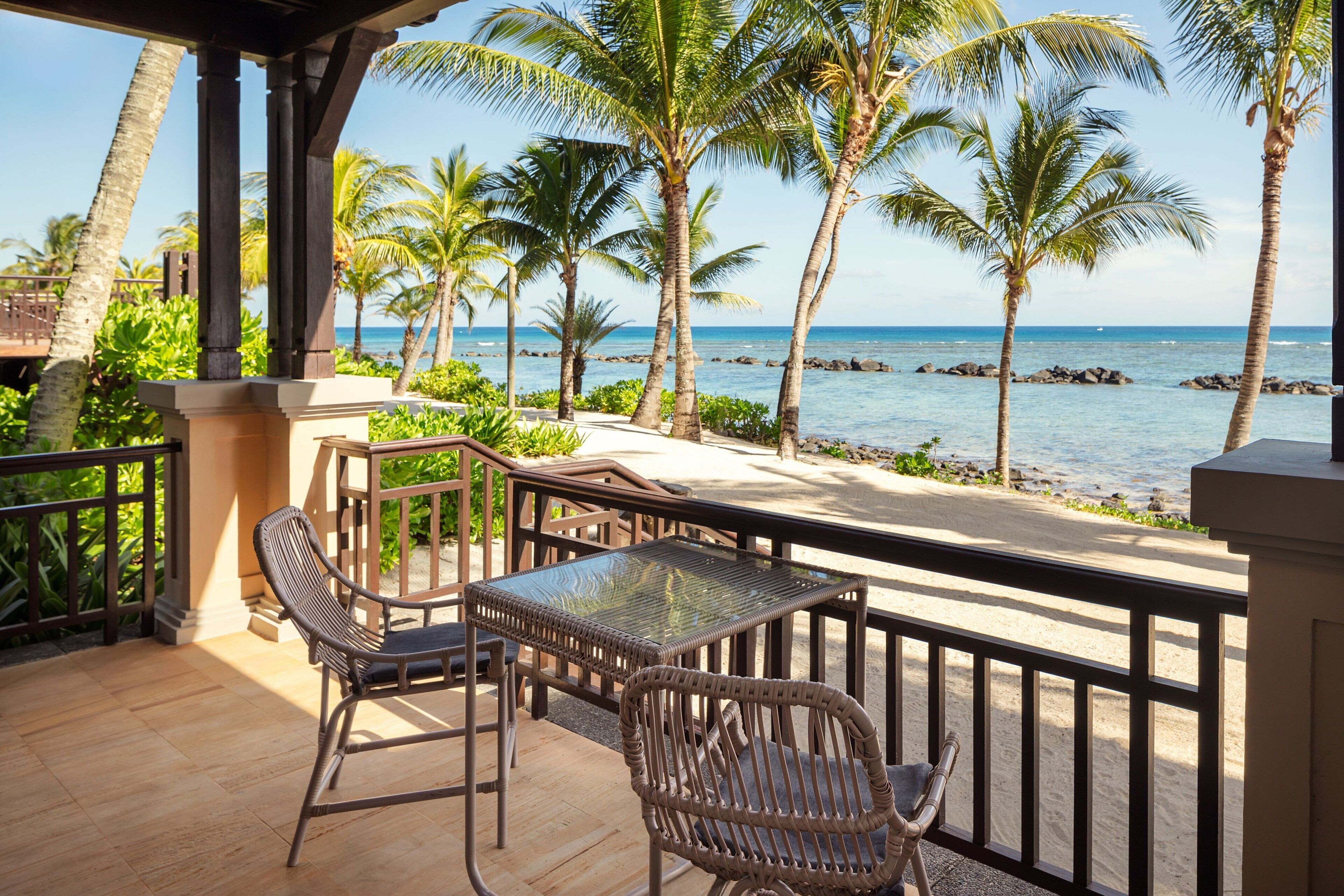A balcony with wicker chairs and a glass table overlooking a beach with palm trees and the ocean.