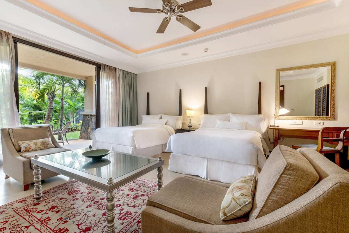Quarto de hotel com duas camas, um sofá, uma mesa de centro de vidro, uma escrivaninha com cadeira e um espelho grande.