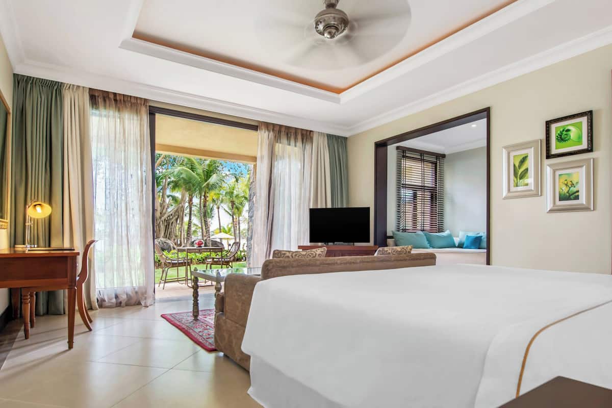 Quarto de hotel com uma cama grande, uma escrivaninha com luminária, televisão e varanda com vista para palmeiras.