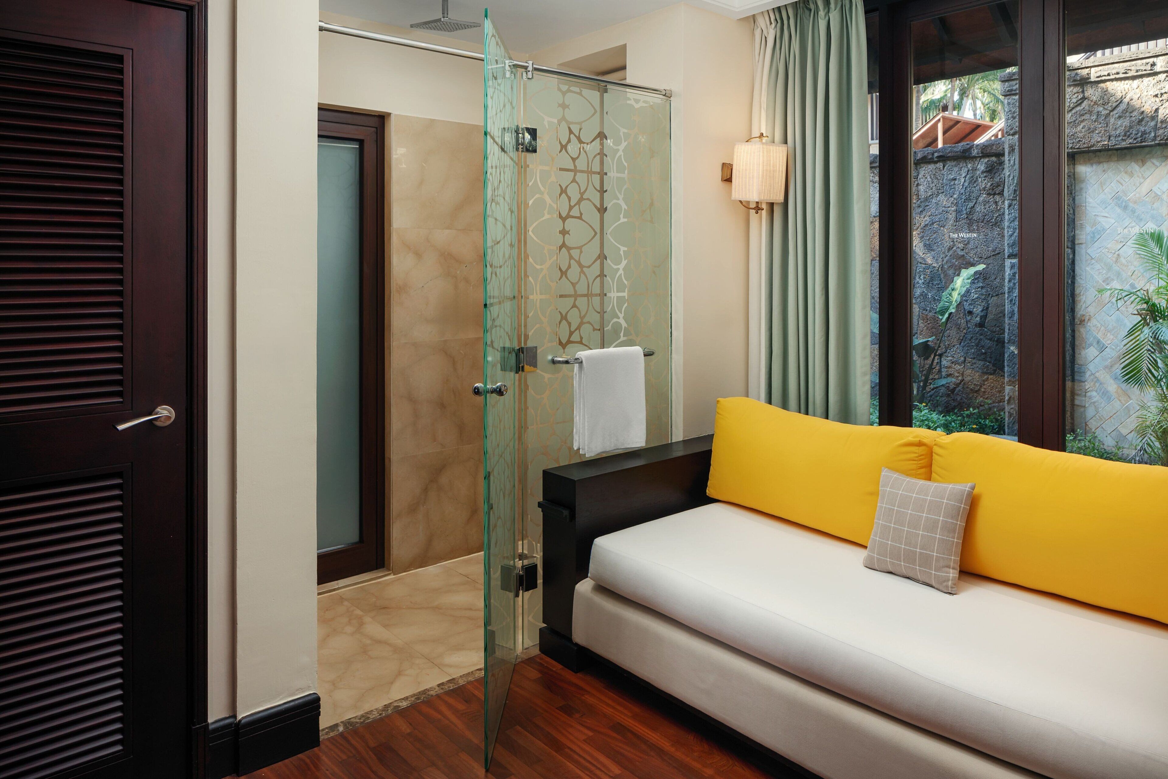 A modern bathroom with a glass shower enclosure, a wooden door, and a yellow sofa with a white cushion.