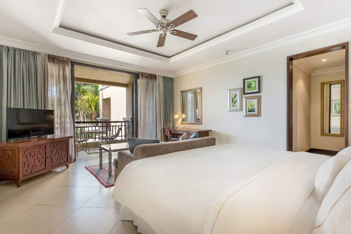 Quarto de hotel com cama, televisão, escrivaninha e varanda com vista para palmeiras.