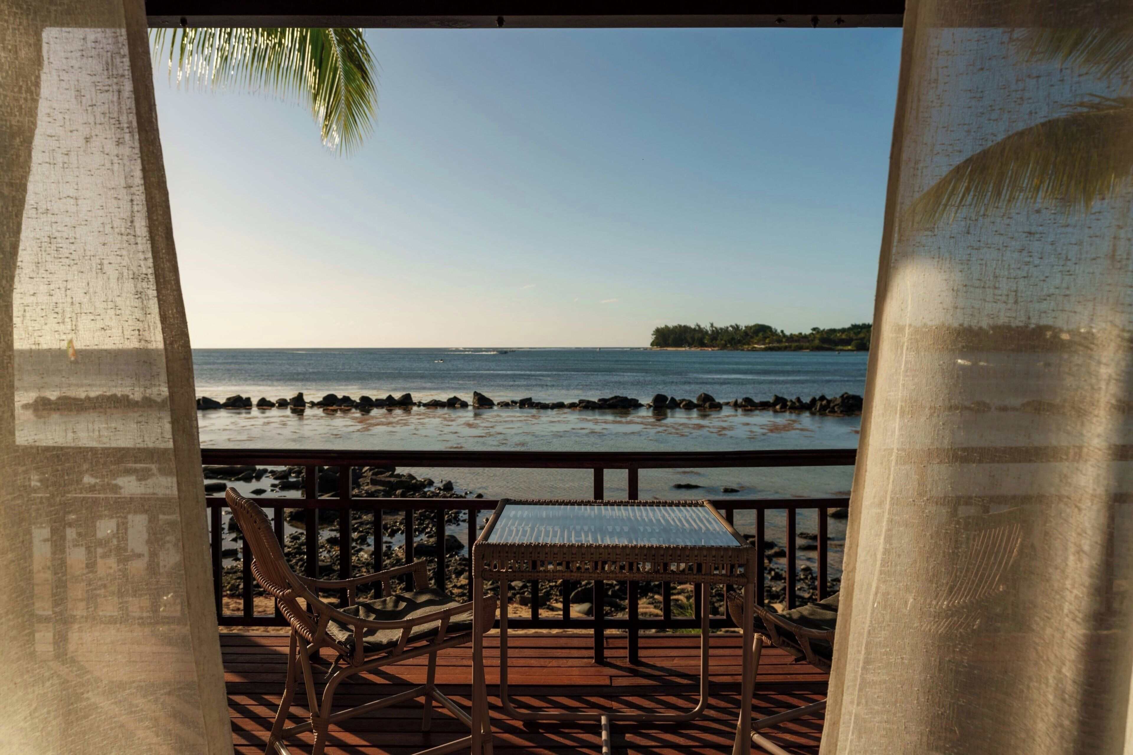 A wooden deck with a table and chairs, overlooking a beach with rocks and a clear sky.