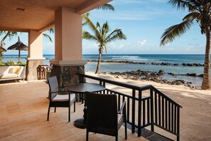 A covered outdoor seating area with a view of the ocean, featuring a stone fireplace, wicker chairs, and a wooden table.