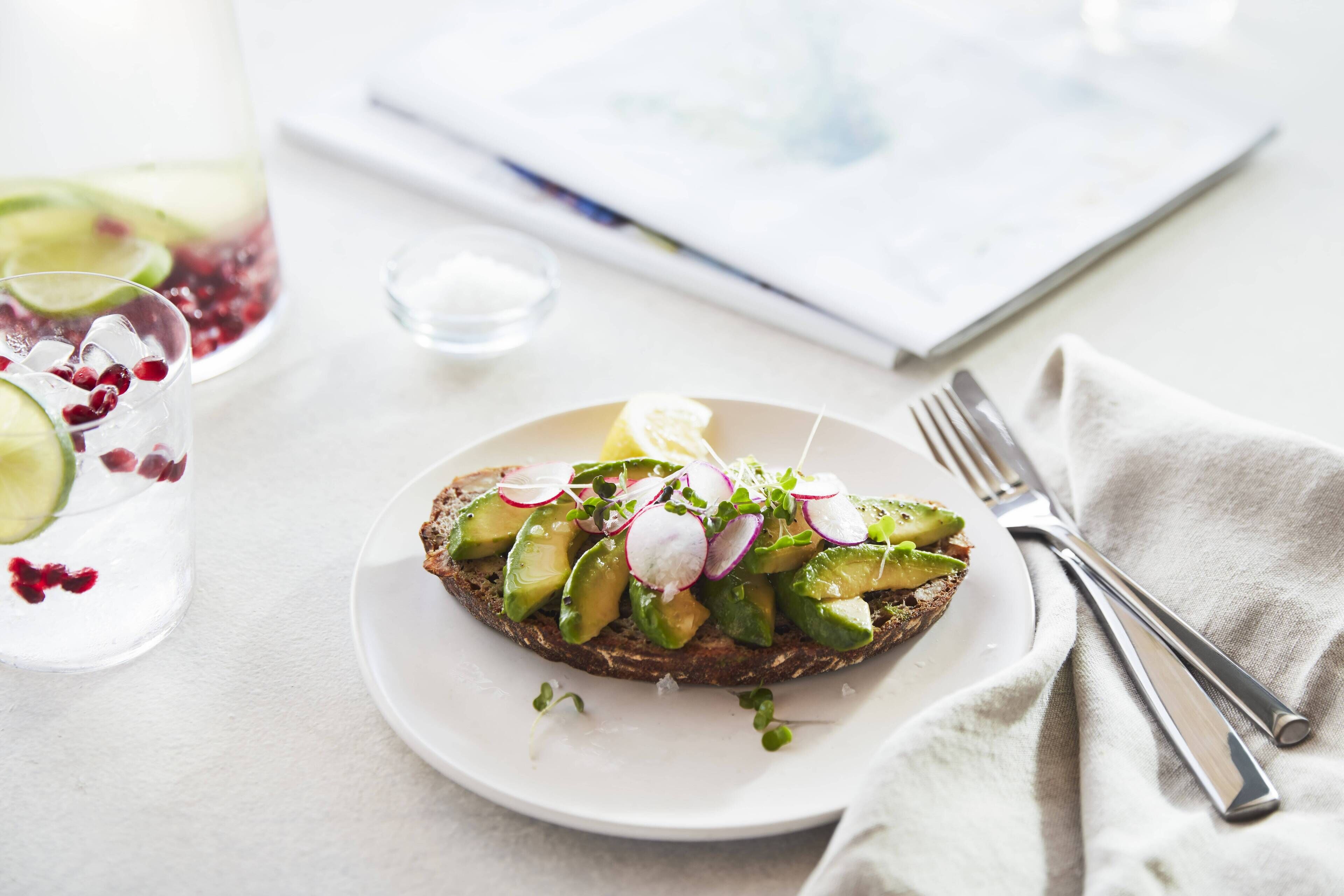 A plate with a slice of toast topped with avocado and radishes, a glass of water with lime and berries, and a fork and knife on a napkin.