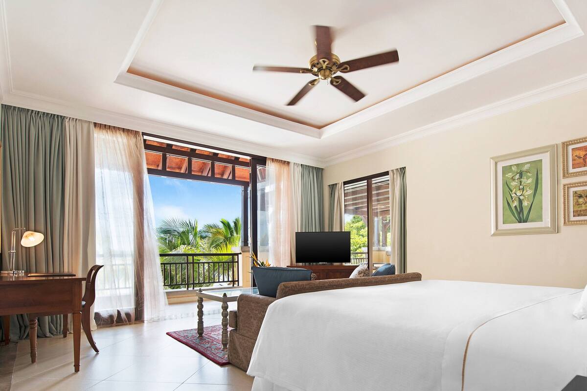 Quarto de hotel com cama, escrivaninha, cadeira, televisão e varanda com vista para palmeiras.
