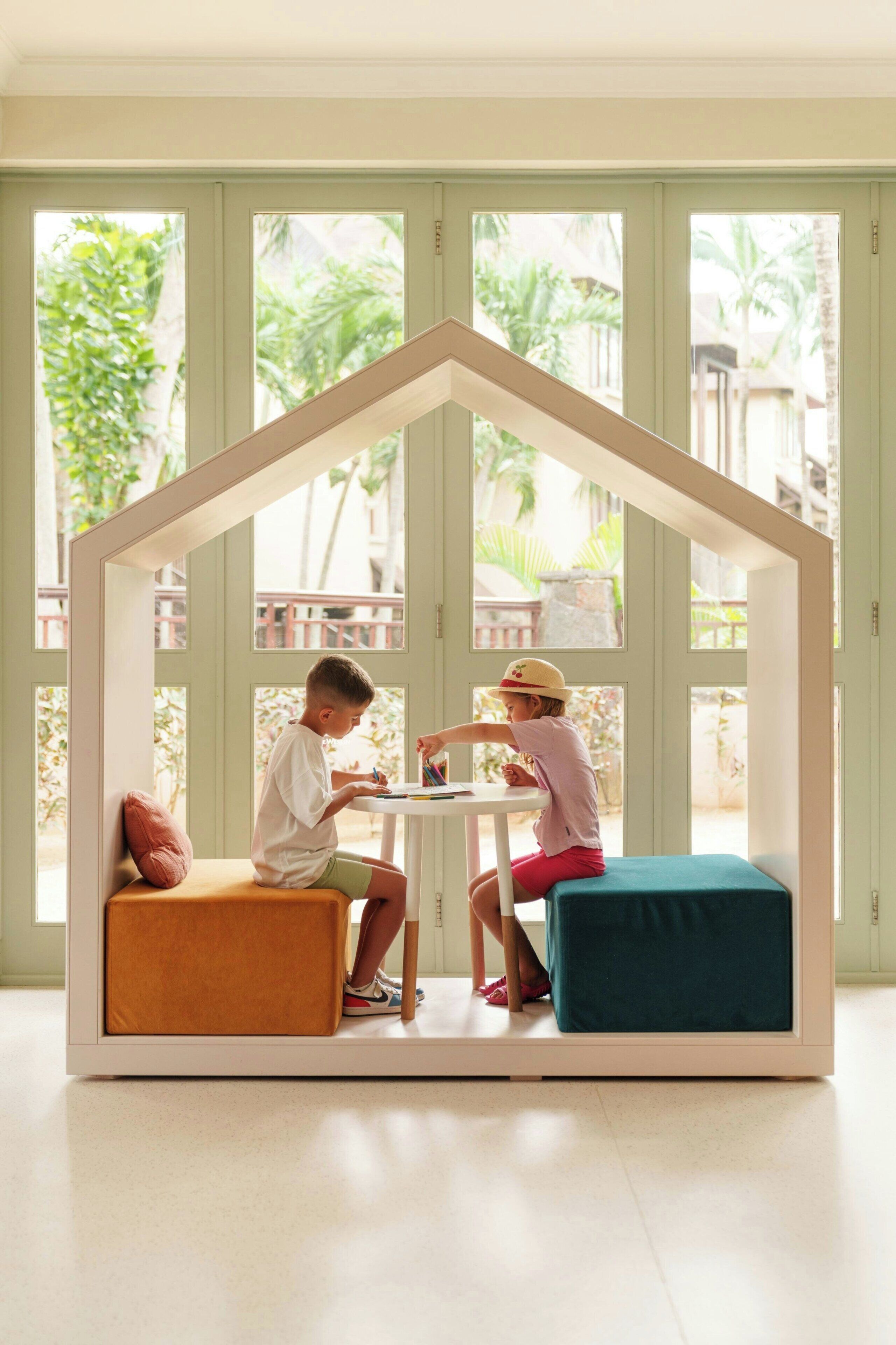 Ein Zimmer mit einem hauseigenen Spielhäuschen in der Mitte, zwei Kinder sitzen an einem Tisch, und große Fenster mit Blick auf grüne Vegetation draußen.